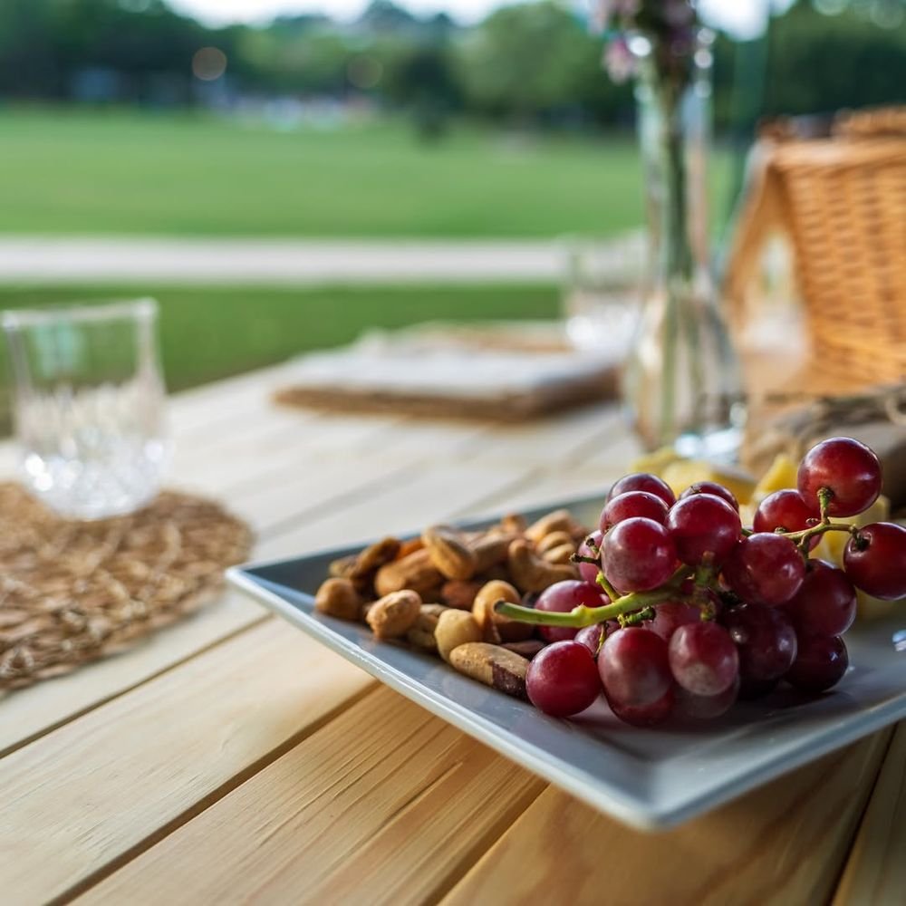 A plate of grapes, cheese and nuts on the pine table with an open green lawn behind.