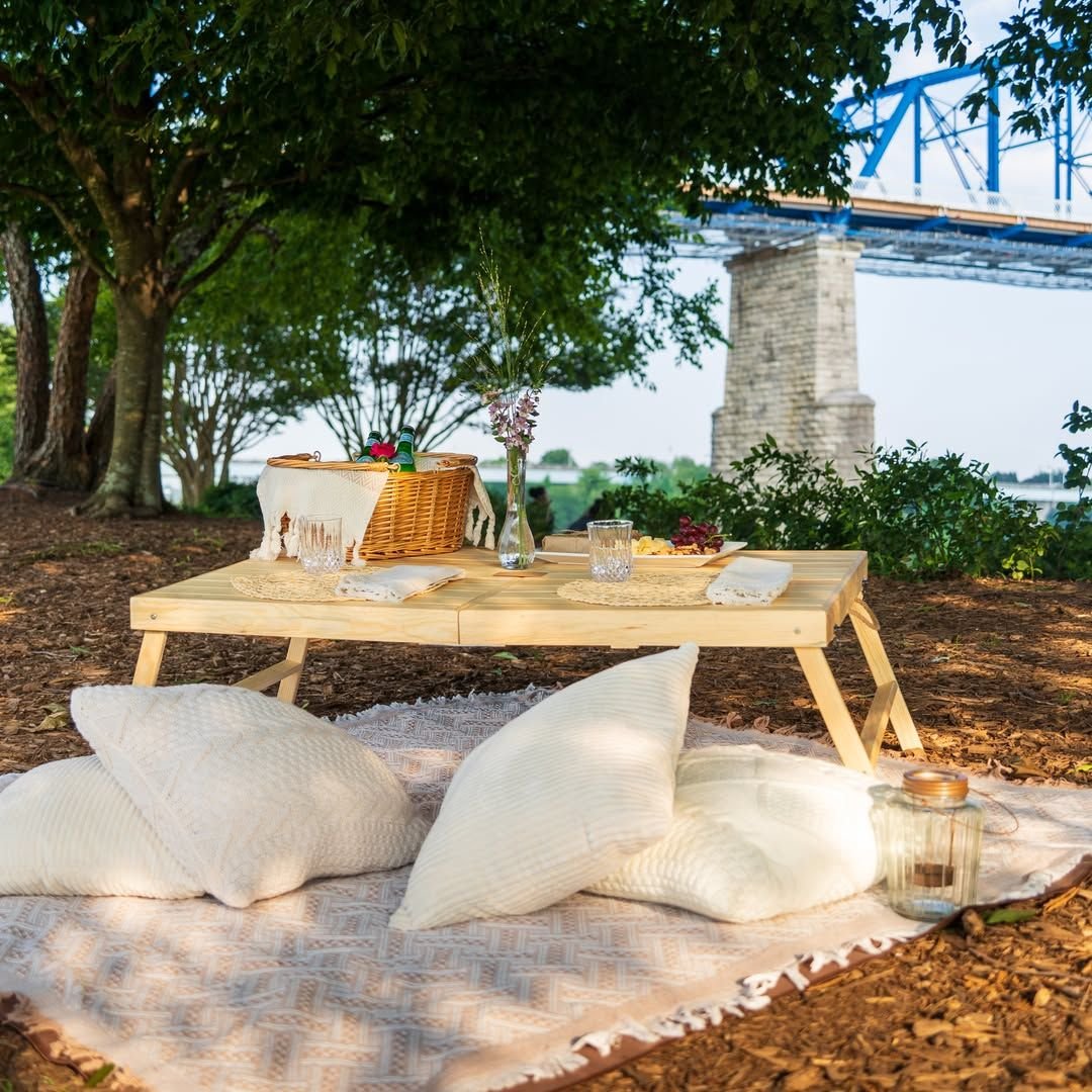A low pine table set with glasses, a bud vase and a basket, on a blanket with cushions under a tree by the river, with the blue bridge behind.